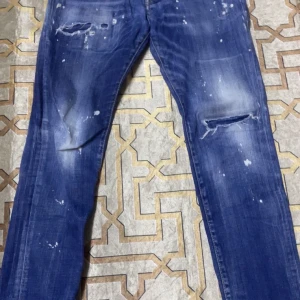 Blå distressed jeans från Dsquared2 - Säljer ett par blå jeans från Dsquared2 med slitningar, färgstänk och hål på benen. Jeansen har raka ben och klassisk femficksdesign. Snygga detaljer på bakfickorna och coolt tvättad look. Perfekta för dig som gillar streetstyle.