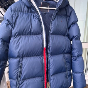 Blå pufferjacka från Tommy Hilfiger - Snygg marinblå pufferjacka från Tommy Hilfiger med huva och dragkedja. Jackan har två fickor med dragkedja och klassiska Hilfiger-detaljer på bröstet och ärmen. Perfekt för kalla dagar och riktigt skön att bära.