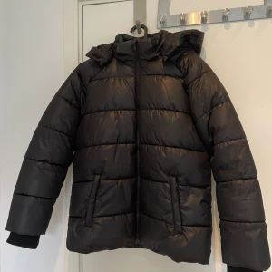 Svart pufferjacka från Kappahl stl 158 - Svart pufferjacka från Kappahl i nyskick med huva och dragkedja framtill. Jackan har quiltad design, två fickor och ribbade muddar vid ärmslut. Tillverkad i 100% polyester, perfekt för kalla dagar. Reflextape baktill för extra synlighet. Stor i storlek mer som 164.