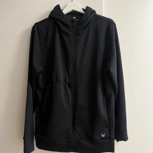 Svart hoodie med dragkedja från SOC - Svart hoodie från SOC i storlek M med hel dragkedja och huva. Tröjan har långa ärmar och en enkel, clean look med liten logga vid fickan. Perfekt för chill eller träning, tillverkad i mjuk polyester. Snygg och basic design.