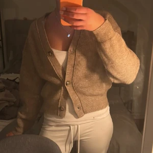 Brun ribbad kofta med knappar - Mysig brun -beige kofta med ribbad struktur och v-ringning. Koftan är croppad och har stora knappar framtill. Perfekt att slänga över ett linne eller t-shirt för en chill look. Långa ärmar och mjukt stickat material.