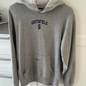 Grå hoodie Southfield med huva - Grå hoodie från Southfield med mörkblå tryck på bröstet och stor ficka framtill. Tröjan har huva med snörning och långa ärmar, perfekt för chill dagar. Mjuk bomullskänsla och klassisk loose passform.