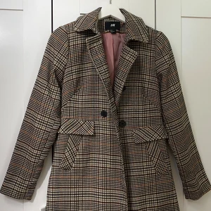 Rutig kappa från H&M i brun och beige - Snygg rutig kappa från H&M i storlek 36. Jackan har klassisk krage, två stora fickor med lock och stängs med svarta knappar. Färgerna går i beige, brunt och svart med inslag av vitt. Fodret är ljusrosa och materialet känns mjukt och bekvämt. Kappa från H&M, som ni ser på bilden så har den inga fläckar eller är skadad. Längden är 80 cm och bredden (från ena axeln till den andra) är 37 cm. Meddela mig gärna för mer bilder eller info!💕