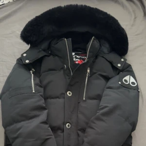 Svart dunjacka från Moose Knuckles - Säljer en svart dunjacka från Moose Knuckles med stor pälskantad huva, silverfärgade dragkedjor och knappar. Jackan har logga på ärmen och flera praktiska fickor. Perfekt för kalla dagar och riktigt snygg streetstil. Pris kan diskuteras.