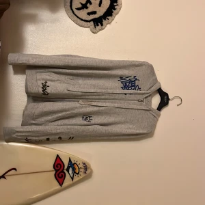 Grå hoodie Zadig & Voltaire med tryck - Grå hoodie från Zadig & Voltaire med coola grafitti-inspirerade tryck och broderier i blått och svart. Tröjan har dragkedja framtill, huva och långa ärmar. Mjuk bomullskänsla och streetig vibe, perfekt för dig som gillar unika detaljer.