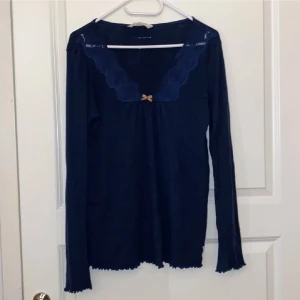 Mörkblå långärmad topp Odd Molly - Supersöt mörkblå långärmad topp från Odd Molly med spetskantad v-ringning och liten beige rosett framtill. Modellen är ribbad och har vågig kant nertill och vid ärmslut. Perfekt för dig som gillar romantiska detaljer och mjuka material.