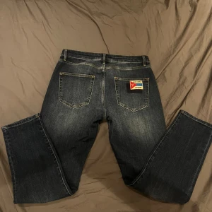 Saint Laurent Université jeans blå - Mörkblå jeans från Saint Laurent med snyggt tvättad finish och kontrastsömmar. Bakfickan har en unik patch med texten 'Saint Laurent Université' i rött, blått och gult. Klassisk femficksmodell med raka ben och metallknapp med logga.