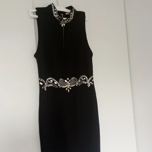 Svart kort klänning med pärldetaljer - Supersnygg svart ärmlös klänning med hög krage och glittrande pärldetaljer vid halsen och midjan. Klänningen har en markerad dragkedja bak och coola. Perfekt för dig som vill sticka ut med en elegant och edgy look.