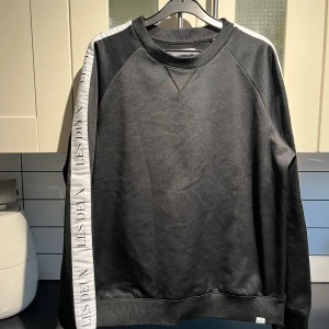 Svart sweatshirt från Les Deux med vit detalj - Svart sweatshirt från Les Deux med vit panel och logga längs ena ärmen. Klassisk rund halsringning och ribbade muddar. Snygg och enkel design som passar perfekt till jeans eller joggers.