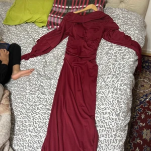 Vinröd långklänning med v-ringning - Säljer en vinröd långklänning med v-ringning och långa ärmar. Klänningen har en snygg drapering över magen och en bred krage som ger en cool look. Materialet är mjukt och följsamt, perfekt för dig som vill sticka ut.