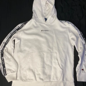 Vit hoodie från Champion med logoband - Vit hoodie från Champion med svart logoband längs ärmarna och broderad Champion-logga på bröstet. Klassisk känguruficka framtill och huva. Mjuk bomullskänsla och ribbade muddar vid ärmslut och midja. Perfekt för en sportig och avslappnad stil.