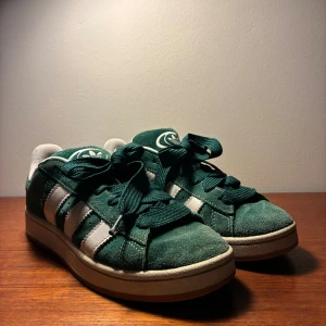 Adidas Campus gröna sneakers mocka - Gröna Adidas Campus sneakers i mocka med vita detaljer och klassiska tre ränder på sidan. Rund tå, platt sula och snörning. Adidas logga på plösen. Medföljer extra skosnören i grönt och vitt. Perfekt för dig som gillar retrostil och vill sticka ut.