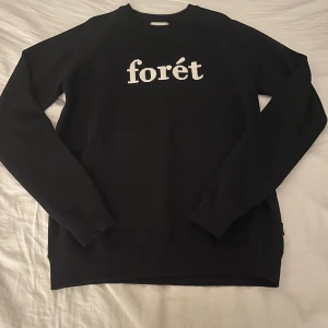 Svart sweatshirt från Forét i bomull - Svart sweatshirt från Forét med vit broderad logga på bröstet. Tillverkad i ekologisk bomull, har rund hals och ribbade muddar vid ärmslut och nederkant. Perfekt för dig som gillar clean och enkel stil.
