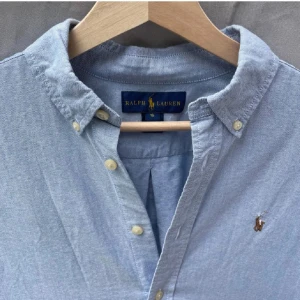 Blå skjorta från Ralph Lauren - Säljer en klassisk ljusblå skjorta från Ralph Lauren i storlek L. Skjortan är i bomull och har en broderad logga på bröstet. Den har långa ärmar och knappar framtill. Perfekt för både vardag och fest! 