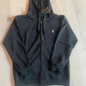 Svart hoodie från Polo Ralph Lauren - Svart hoodie från Polo Ralph Lauren med dragkedja och huva. Klassisk broderad logga på bröstet och snörning i huvan. Mjuk bomullskvalitet och ribbade muddar vid ärmslut och nederkant. Perfekt för en chill och stilren look.