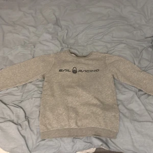 Grå Sail Racing sweatshirt - Snygg grå sweatshirt från Sail Racing med svart logga på bröstet och liten logga på baksidan. Den har en riktigt snygg look och passar både att bara ha på skolan eller ha den vid finare tillfällen. Priset går att diskuteras