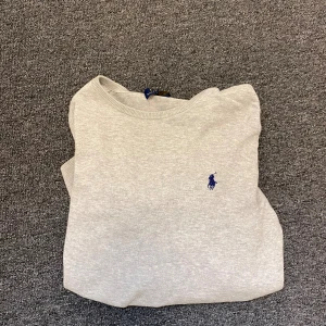 Ljusgrå tröja från Polo Ralph Lauren - Snygg ljusgrå tröja från Polo Ralph Lauren med klassisk rund hals och broderad blå logga på bröstet. Tröjan är långärmad och har ribbade muddar vid ärmslut och nederkant. Perfekt för en clean och stilren look.