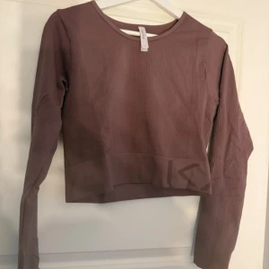 Ribbad brun/rosa croppad topp från Kay/Day - Säljer en brun ribbad långärmad topp från Kay/Day. Toppen är croppad med diskret tryck längst ner och har en rund halsringning. Materialet är mjukt och stretchigt, perfekt för en chill look. Passformen är tight och den har en snygg, enkel design. Helt oanvänd