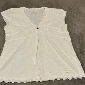 Vit blus med spets från Odd Molly - Säljer en vit blus från Odd Molly med v-ringning, korta ärmar och dekorativ spets längs halsringning och nederkant. Blusen har en liten knapp framtill och broderad logga i nacken. Perfekt för dig som gillar detaljer och en feminin stil.