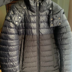 Svart pufferjacka från BOSS med huva - Snygg svart pufferjacka från BOSS med snygg design, hög krage och avtagbar huva. Jackan har dragkedja framtill, tryckknappar på axlarna och diskret BOSS-logga på ärmen. Insidan är fodrad i guldbrun färg för extra stilpoäng. Använd men i mycket gott skick. 