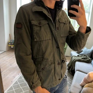 Olivgrön parkajacka från Marc O'Polo - Säljer en olivgrön parkajacka från Marc O'Polo med fyra stora fickor framtill, knappar och dragkedja. Jackan har klassisk krage, patch på ärmen och är tillverkad i bomull. Perfekt för dig som gillar en avslappnad och tidlös stil.