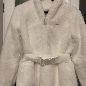 Vit teddyjacka från Aim'n med bälte - Supermjuk och fluffig vit teddyjacka från Aim'n med dragkedja framtill och ett brett bälte i midjan. Jackan har lång ärm och en riktigt cozy vibe, perfekt för kyliga dagar. Snygg och stilren design som sticker ut.
