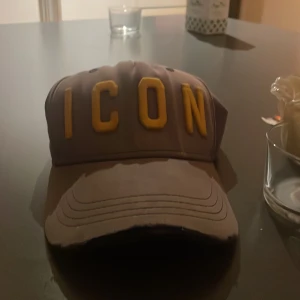 Grå Dsquared2 ICON keps med gul text - Snygg grå keps från Dsquared2 med stor gul 3D-text 'ICON' framtill och broderad Dsquared2-logga baktill. Justerbar rem med metallspänne för perfekt passform. Kepsen är tillverkad i bomull och har klassisk böjd skärm.