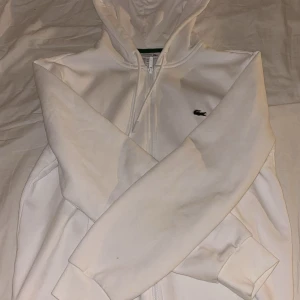 Vit hoodie från Lacoste med dragkedja - Säljer en stilren vit hoodie från Lacoste med dragkedja framtill, huva med snörning och klassisk krokodillogga på bröstet. Tröjan har långa ärmar och två fickor framtill. Perfekt för en clean och sportig look.