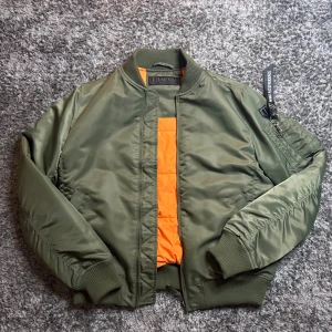 Olivgrön bomberjacka från Jackan - Snygg olivgrön bomberjacka från Jackan med orange quiltat foder. Jackan har ribbade muddar, dragkedja, fickor med tryckknapp och en ärmficka med svart tag. Perfekt för dig som vill ha en klassisk streetstil.