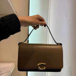 Coach bandit shoulder bag - Säljer en stilren brun axelväska från Coach i slätt  kalvskinn med justerbar rem och snyggt guldfärgat C-spänne framtill. Väskan har en rektangulär form och minimalistisk design som ger en clean look. Perfekt för dig som gillar klassiska accessoarer med en modern twist. Nyskick! Nypris 6895kr