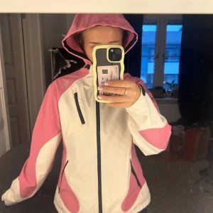 Skidjacka från Bergans - Snygg och fodrad skidjacka från Bergans i rosa och vitt med huva och svarta dragkedjor. Jackan har tre fickor med dragkedja och sportig design. Perfekt för kalla dagar i backen och har en skön passform med långa ärmar. Storlek L men passar mig som vanligtvis bär S