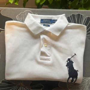 Vit pikétröja Polo Ralph Lauren M - Vit pikétröja från Polo Ralph Lauren i slim fit-modell. Klassisk krage, knäppning med två knappar och stor mörkblå broderad logga på bröstet samt siffra 3 på ärmen. Tillverkad i bomull, perfekt för en clean och sportig stil.