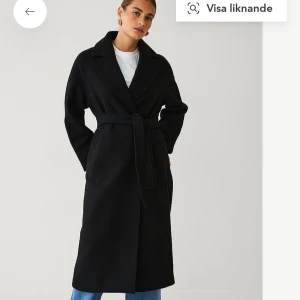 Svart lång kappa med bälte - Stilren svart kappa från Ellos med klassisk krage och knytskärp i midjan. Lång modell med raka ärmar och snyggt fall. Aldrig använd!! Nypris 1199kr, 30% ull