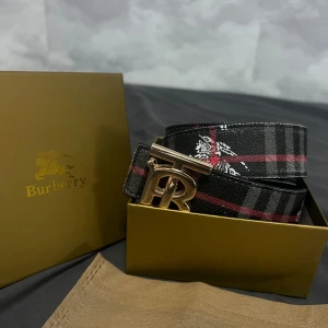 Burberry bälte - Snyggt Burberry-bälte i svart skinn med klassiskt rutig design i rött, vitt och grått. Stort guldigt 'TB'-spänne framtill. Kommer i original ask och dustbag. Perfekt accessoar för att lyfta din outfit med lyxig känsla.