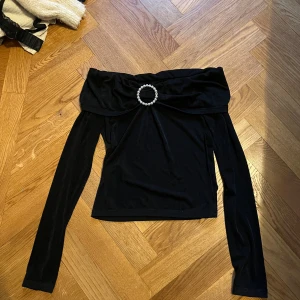 Svart offshoulder topp med pärldetalj - Snygg svart offshoulder topp med lång ärm och en bred krage som vilar över axlarna. Framtill finns en rund pärldetalj som ger extra bling. Materialet är mjukt och har en lätt glans, perfekt för att sticka ut. Passformen är normal och toppen är croppad i modellen.