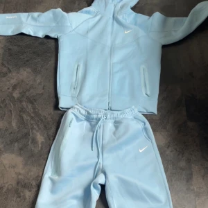 Ljusblå Nike NOCTA hoodie med dragkedja - Säljer en ljusblå hoodie från Nike NOCTA med hel dragkedja och huva. Tröjan har långa ärmar, ribbade muddar och diskreta vita loggor på bröstet och ärmarna. Perfekt för dig som gillar streetwear och sportig stil.