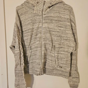 Gråmelerad hoodie med spets från Superdry - Gråmelerad hoodie från Superdry med dragkedja, huva och snygga spetsdetaljer längs ärmar och huva. Tröjan har två fickor med dragkedja och är långärmad. Liten fläck  nere på högra sidan men annars fint skick