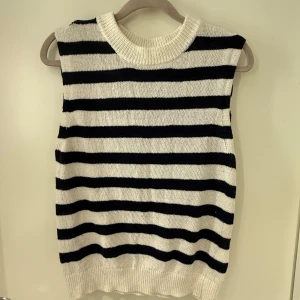 Randig stickad väst i svart och vitt - Snygg stickad väst med breda svarta och vita ränder. Modellen är ärmlös med rund hals och ribbad kant nedtill och vid halsen. Perfekt att styla över en skjorta eller t-shirt för en trendig look.