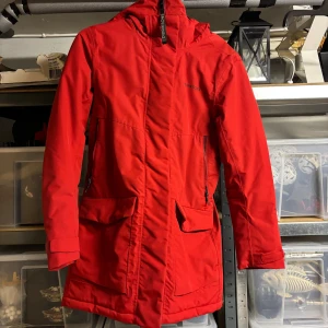 Röd parkajacka från Didriksons - Säljer en röd parkajacka från Didriksons med huva, dragkedja och stora fickor framtill. Jackan är quiltad på insidan och har justerbar midja baktill. Materialet är polyamid och polyester, vilket gör den både varm och 100% vattentät. Perfekt för kalla och regniga dagar.