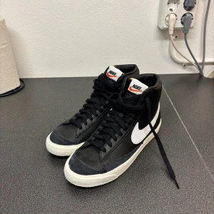 Nike Blazer Mid svart/vit sneakers - Nike Blazer Mid sneakers i svart och vitt med klassisk retrodesign. Skorna har högt skaft, snörning och en vit sula med rund tå. Materialet är skinn och syntet, med tydlig vit Nike-logga på sidan och bak. Perfekt för dig som gillar streetstyle och sportig look.