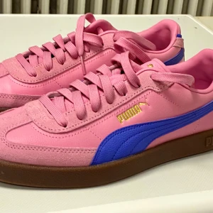 Puma Club II Era rosa/blå sneakers - Säljer ett par Puma Club II Era sneakers i storlek 40. Skorna är ljusrosa med blå sidodetalj och guldiga Puma-loggor. Ovandelen är i skinn och mocka, med bruna gummisulor och klassisk snörning. Perfekt för dig som gillar färgstarka och unika sneakers.