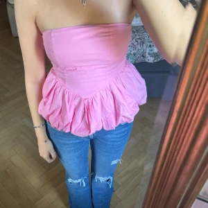 Rosa volang tubtopp - Säljer en söt rosa tubtopp med volang nedtill. Toppen är axelbandslös och har en bandeauformad överdel som sitter tight mot kroppen. Materialet känns som bomull och toppen har en feminin och lekfull look som verkligen sticker ut.