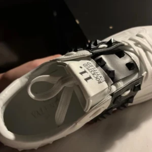 Valentino Rockstud vita sneakers - Snygga vita Valentino Rockstud sneakers med svarta nitar och bred svart rem över vristen. Skorna har snörning och är tillverkade i skinn med coola detaljer som ger en edgy vibe. Perfekta för dig som vill sticka ut med exklusiva och trendiga sneakers.