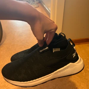 Svarta slip-on sneakers från Reebok - Säljer ett par svarta slip-on sneakers från Reebok med vit sula. Skorna har stickad ovandel i syntetmaterial och elastisk passform utan snörning. Reebok-logga på sidan och hälflik för enkel påtagning. Storlek 38.5