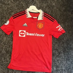 Manchester United Antony 21 matchtröja - Säljer en officiell Manchester United fotbollströja från Adidas med Antony och nummer 21 på ryggen. Tröjan är röd med svarta detaljer, vit krage med röd grafik, Premier League-märke på ärmen och sponsorer tryckta på bröst och ärm. Materialet är lätt och andas, perfekt för match eller träning. 