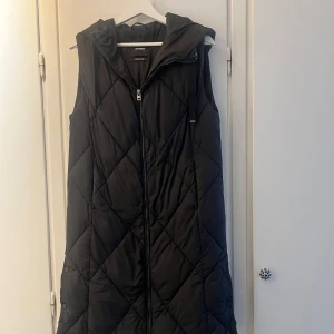 Svart lång  dunväst från Pull and Bear - Svart lång dunväst från Diamond med huva och dragkedja framtill. Västen har fickor på sidan och knappar längst ner på sida 