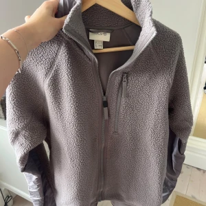 Grå teddyjacka från Move, storlek S - Mysig grå teddyjacka från Move med hel dragkedja och hög krage. Jackan har en bröstficka med dragkedja och detaljer i blankt tyg på insidan av ärmarna. Perfekt för kyliga dagar och har en skön, loose passform.