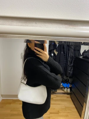 Vit axelväska i croco-imitation - Säljer en stilren vit axelväska med croco-mönstrad yta och dragkedja upptill. Väskan har en enkel, avlång form och ett justerbart axelband. Perfekt för dig som gillar trendiga accessoarer med lite edge.