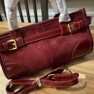 Vinröd mocka axelväska med detaljer - Snygg vinröd axelväska i mocka med skinndetaljer och guldfärgade spännen. Väskan har ett avlångt format, justerbar axelrem och dragkedja upptill med guldfärgad zip-tag. Perfekt för dig som gillar stilrena accessoarer med lite edge.