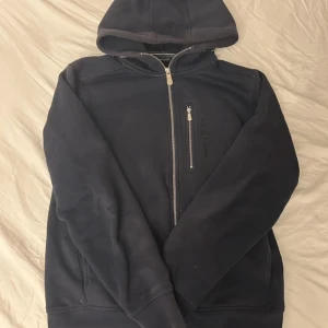 Marinblå sail racing - Marinblå hoodiejacka från SAIL RACING med hel dragkedja och extra bröstficka med zip. Jackan har en stor huva, ribbade muddar och broderad logga på bröstet. Perfekt för chill dagar och streetwear-stil. Priset kan diskuteras!